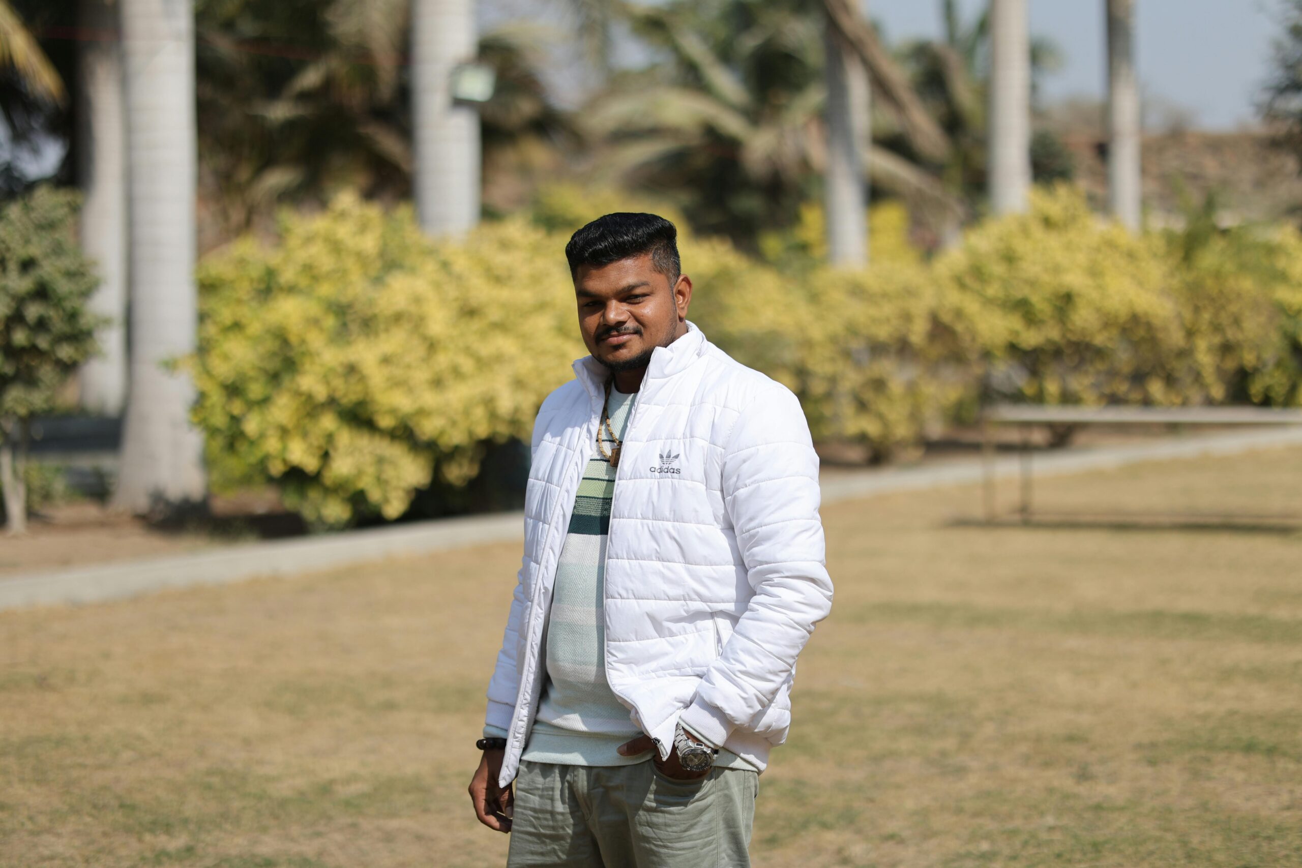 A man in a white jacket stands confidently in a sunny outdoor park setting.